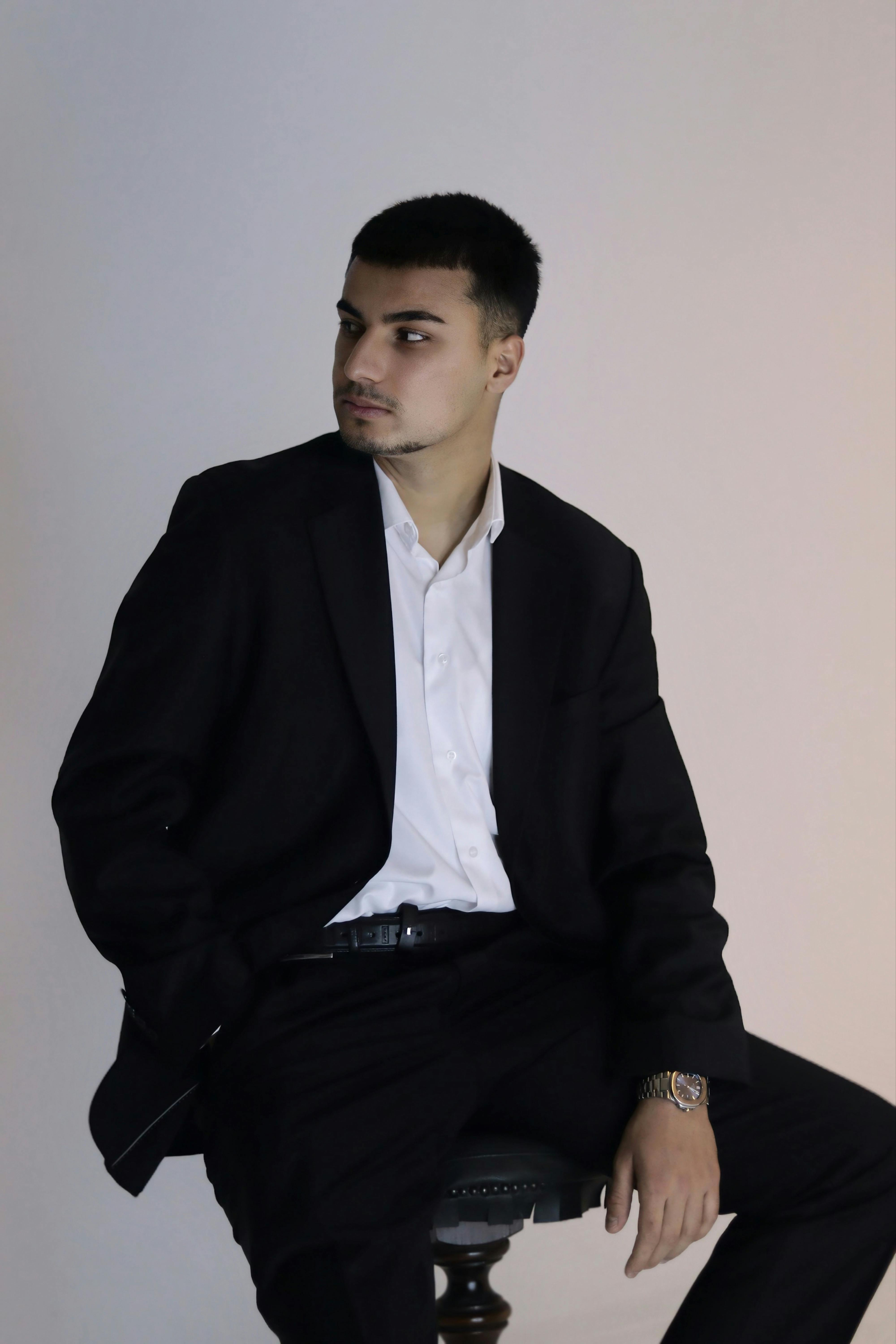 무료 검은색 정장을 입은 젊은 남성이 앉아서 옆을 바라보고 있는 모습의 초상화. 전문적이고 우아한 분위기를 자아낸다. 스톡 사진