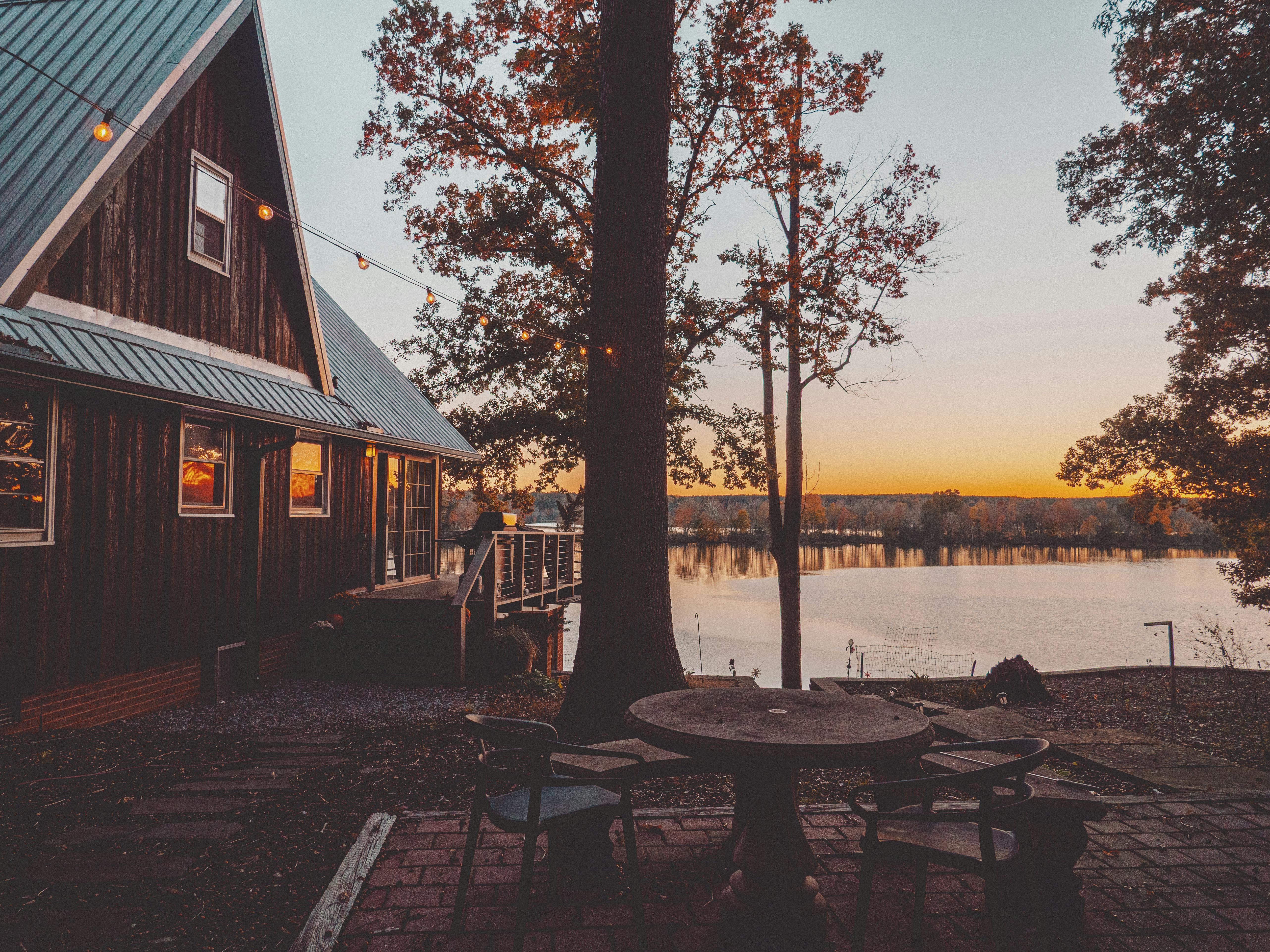 A cozy lakeside house at sunset, adorned with fairy lights, overlooking a serene body of water.