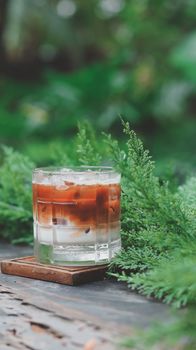 Glass of iced coffee on wooden table surrounded by vibrant green foliage.