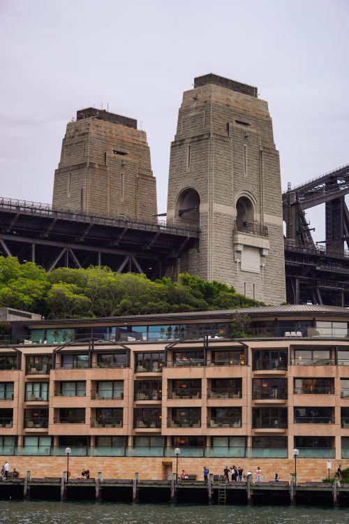 Free View of Sydney Harbour Bridge Pylon with waterfront buildings below, captured on a cloudy day. Stock Photo
