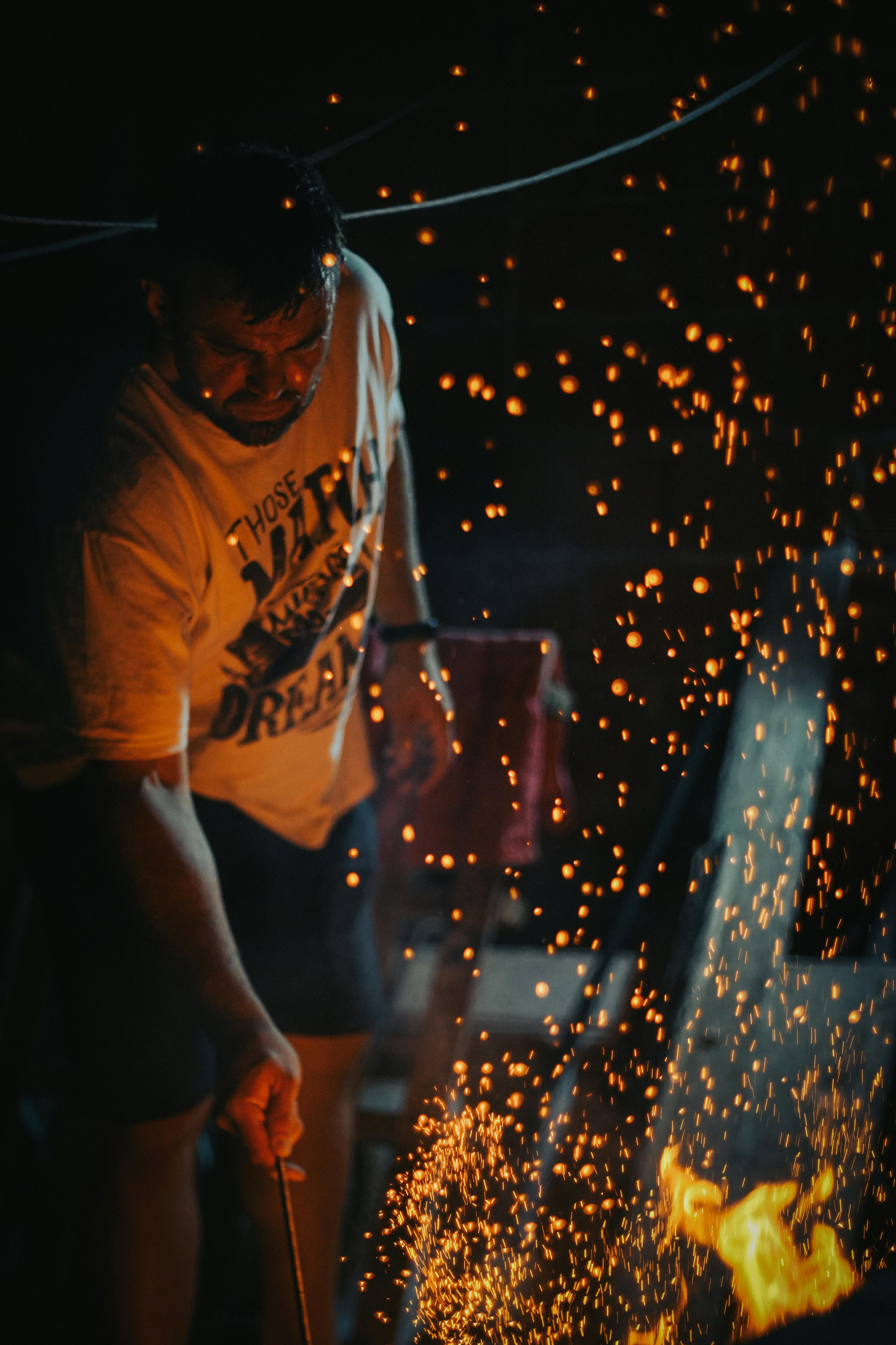 Adult man grilling at night, surrounded by vivid orange sparks and fire in an outdoor setting.