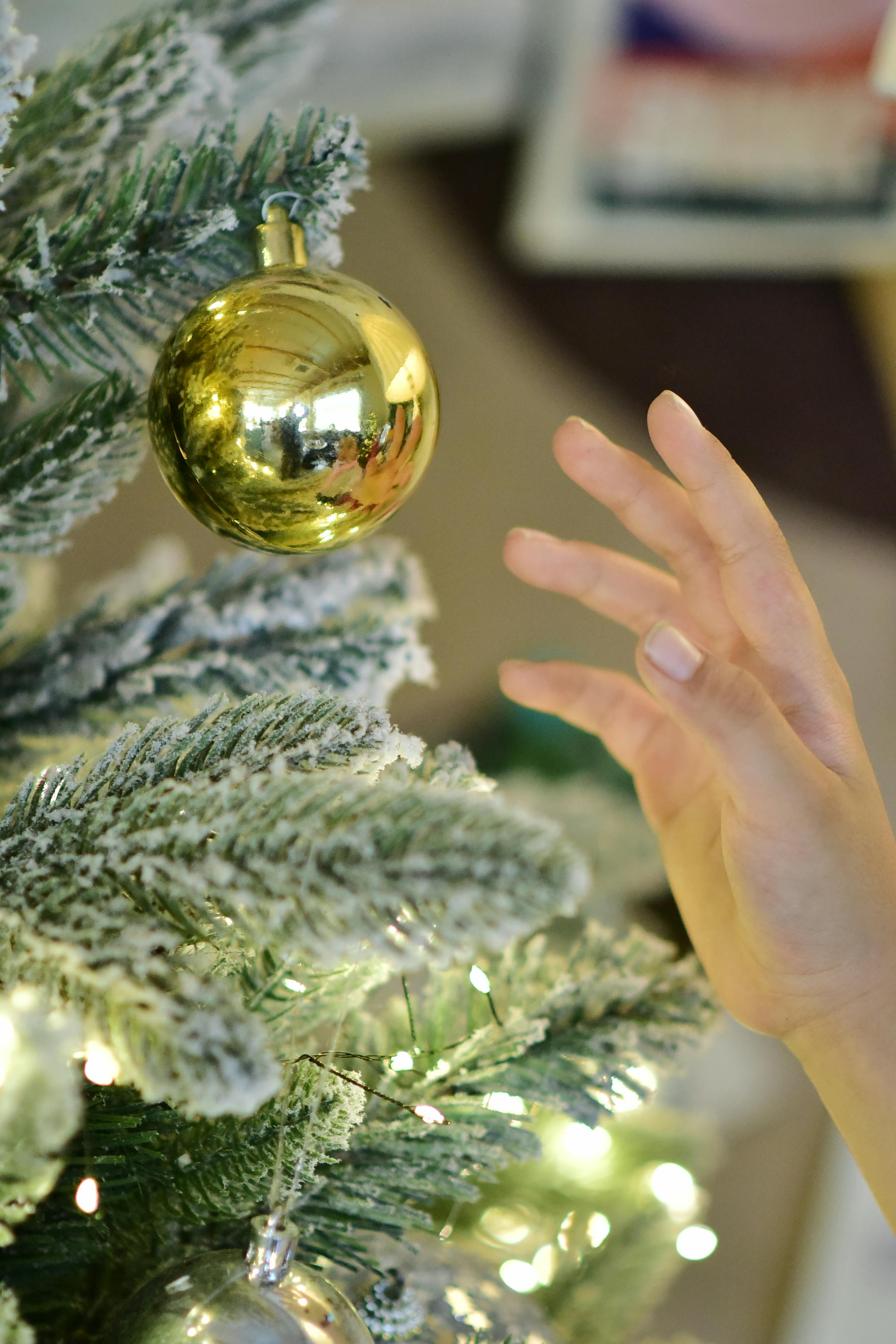 Close-up of a hand reaching to hang a golden bauble on a snowy Christmas tree.