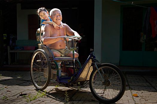 Elderly man and cheerful child enjoying a tricycle ride outdoors in sunlight.