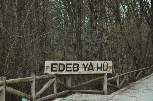 A rustic wooden sign with the text 'Edeb Yâ Hû' in a serene forest setting during fall.