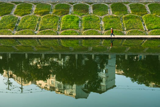 A man walks along a riverside path with urban reflections on calm water.