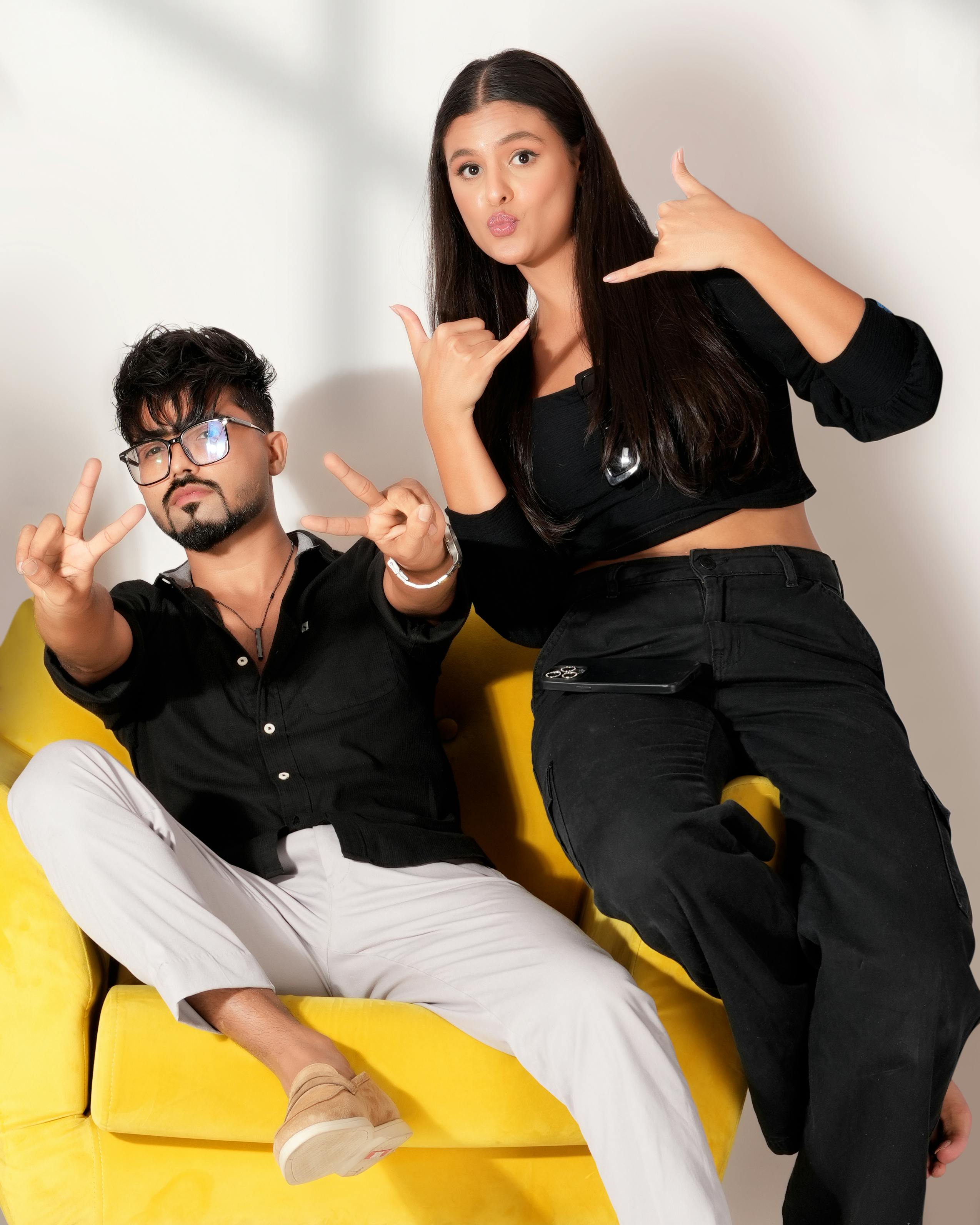 Two friends in black outfits posing playfully on a bright yellow sofa indoors.