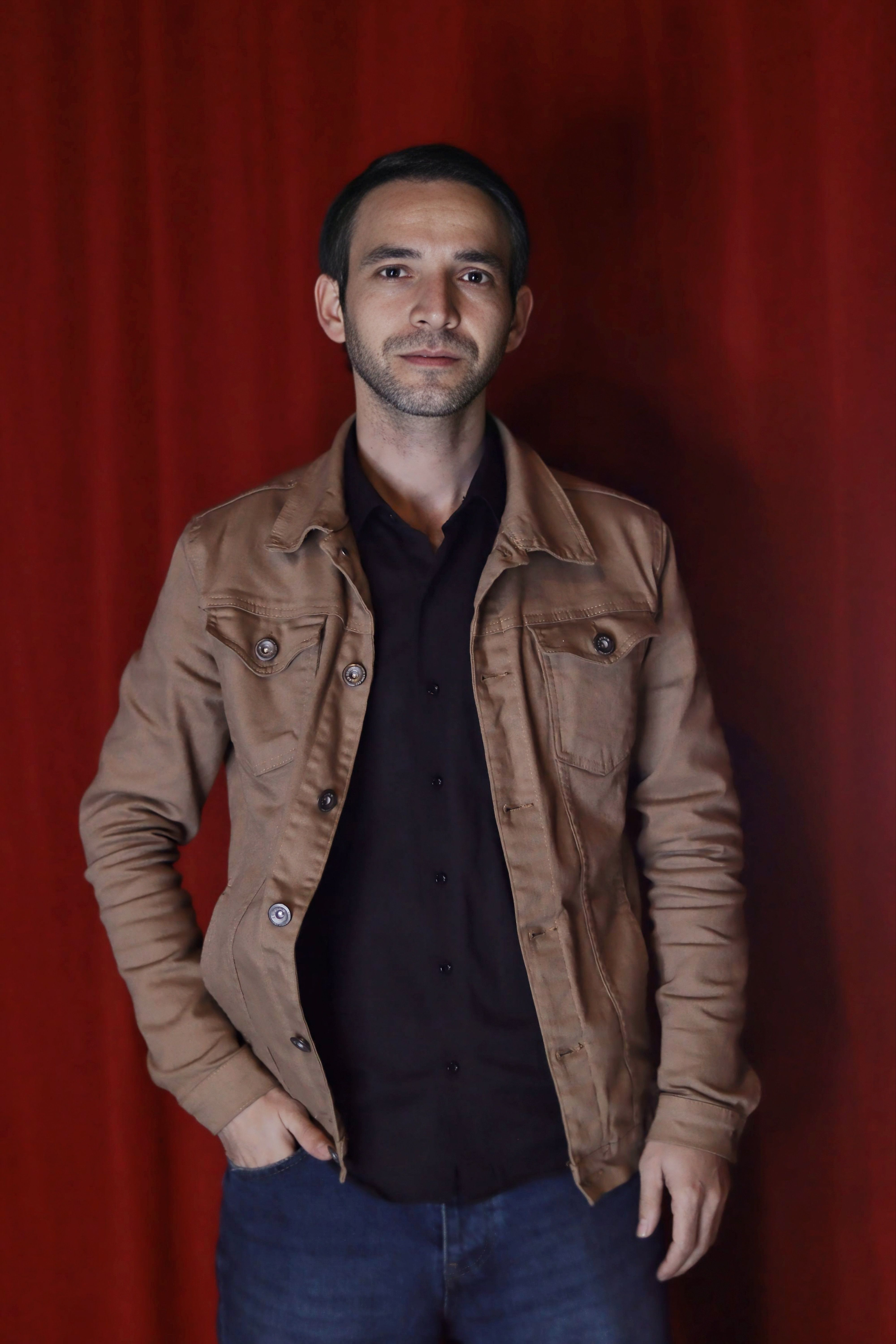 Casual portrait of an adult man in a brown jacket against a vibrant red curtain backdrop.
