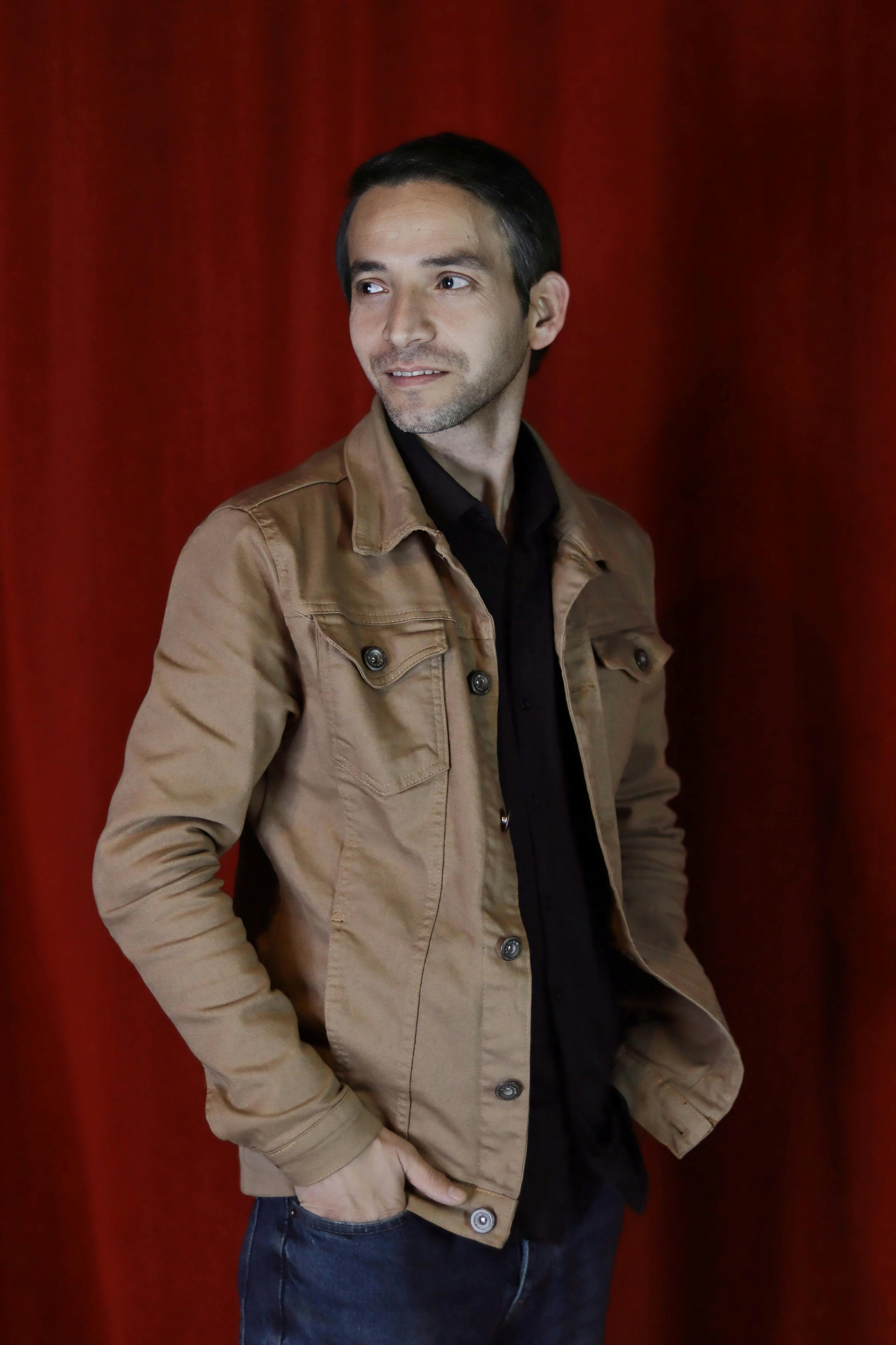 A young man in casual attire standing in front of a vibrant red curtain indoors.