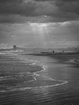 Black and white beach scene with sun rays breaking through clouds over a calm shoreline.