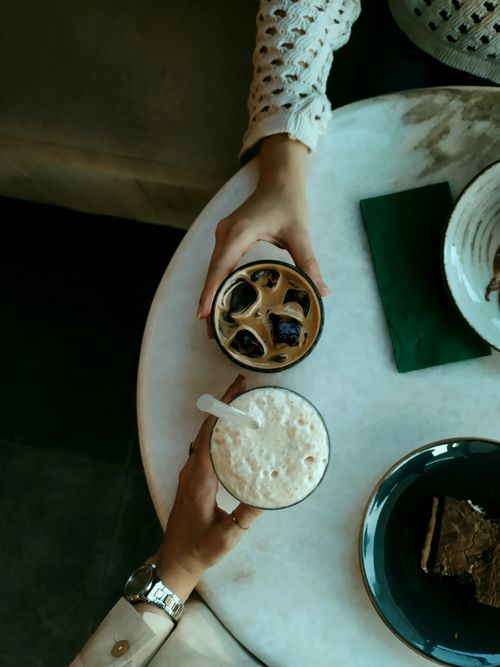 Free Two hands holding iced coffee drinks over a marble table with cake slices, ideal for lifestyle blogs. Stock Photo
