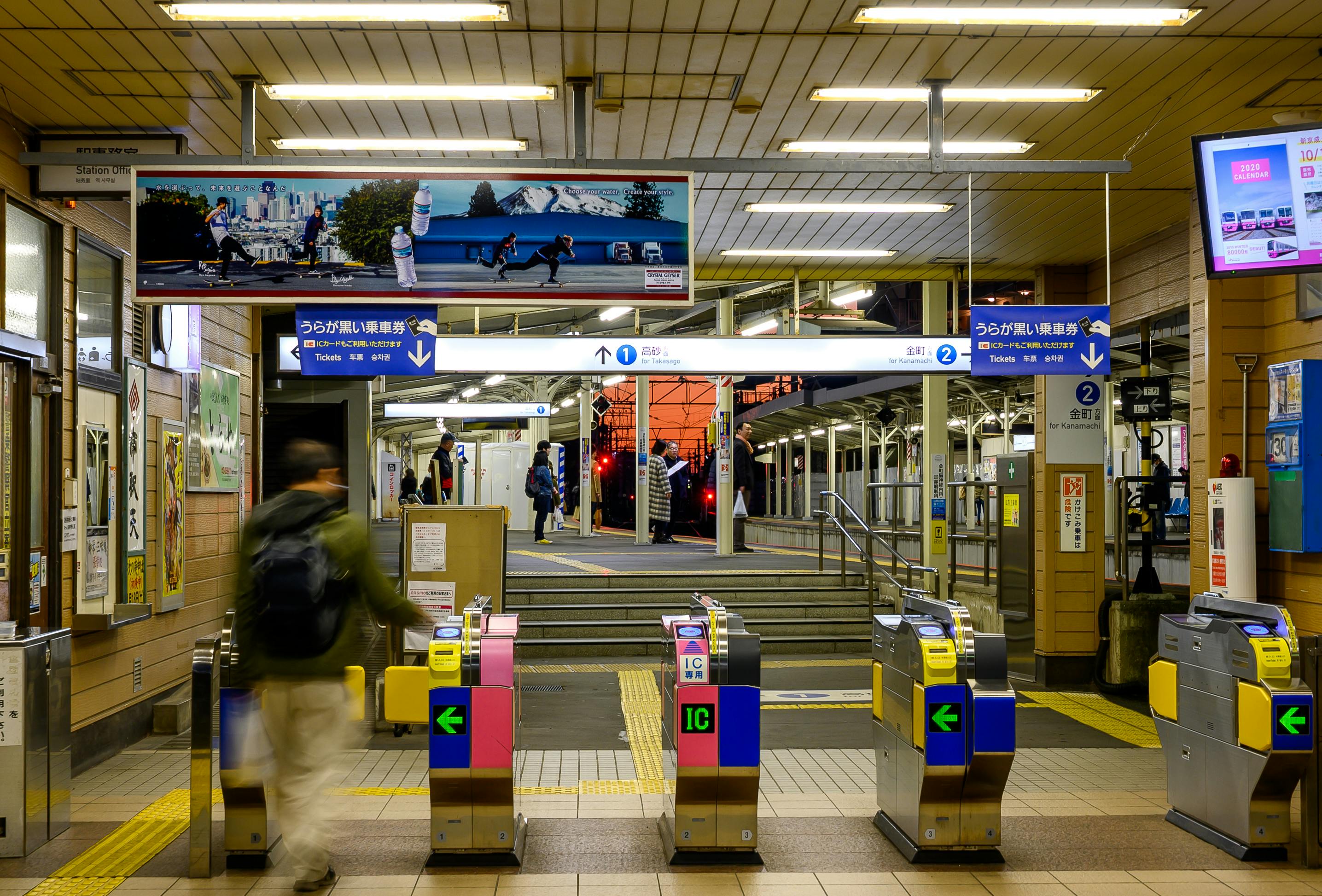 A busy evening scene at a Japanese train station featuring ticket gates and passengers entering the platform area.