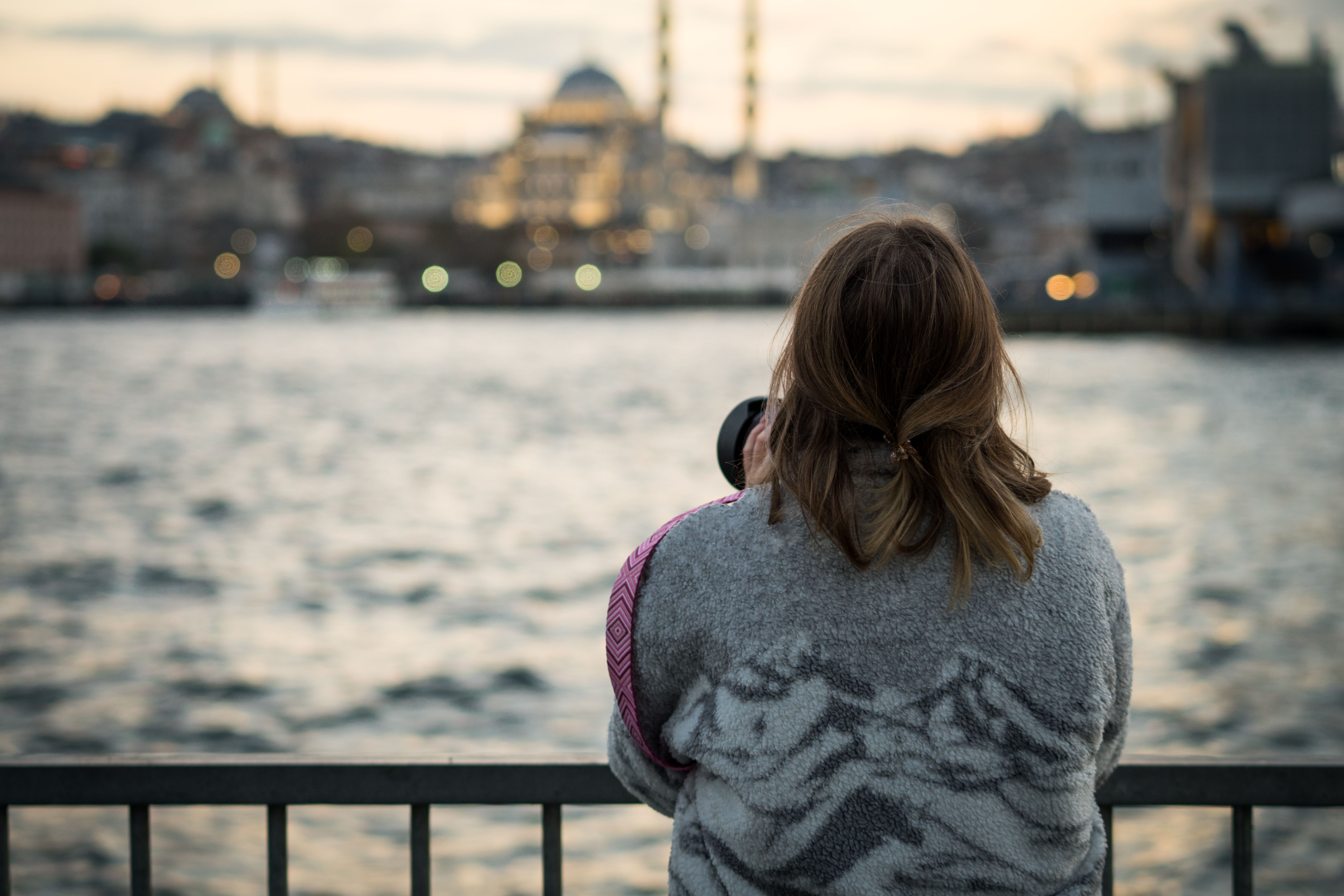 Woman photographs a mosque at sunset, standing by the water in Istanbul.