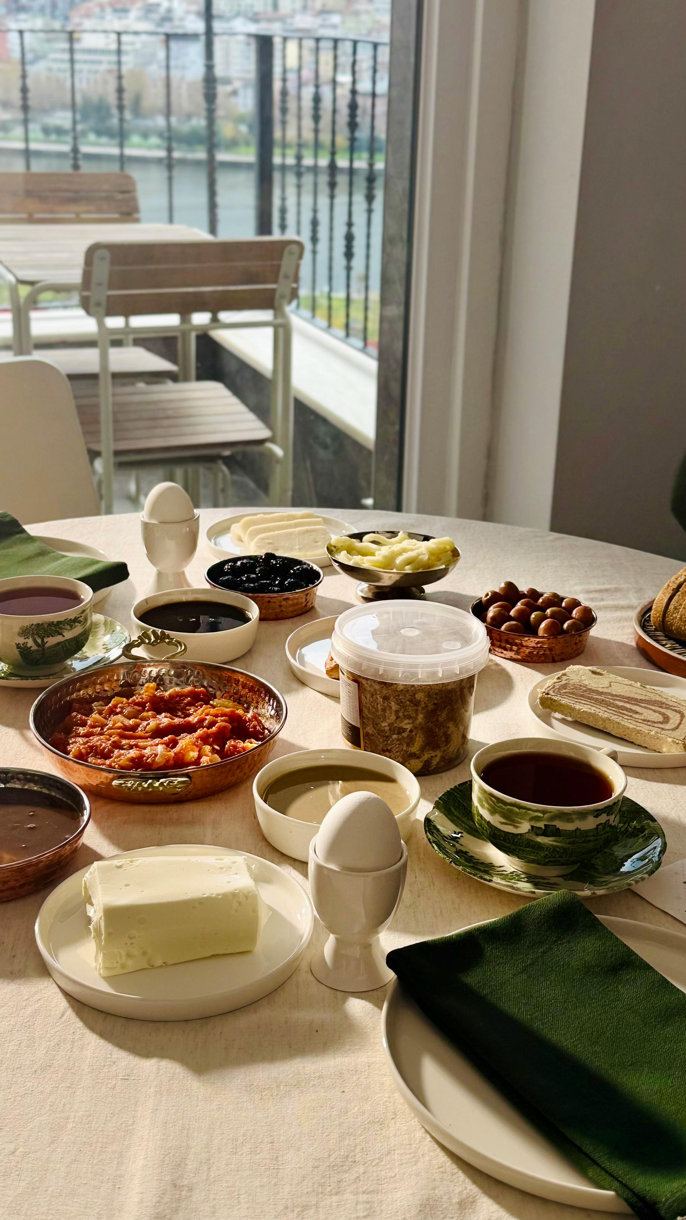 A diverse breakfast spread with eggs, cheese, jams, and tea on a sunlit table by a window.