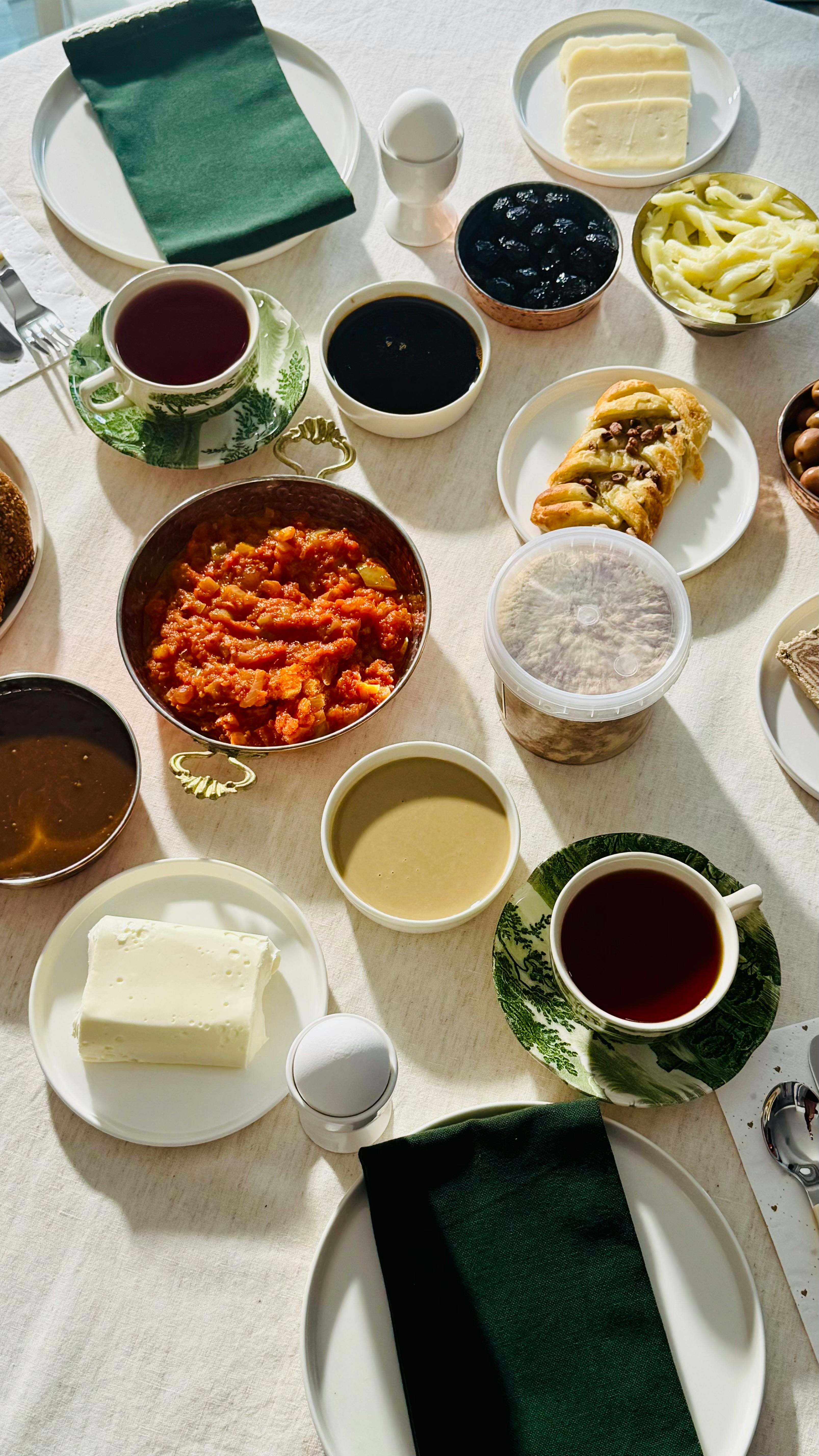 A diverse breakfast table set with tea, spreads, pastries, and desserts on a sunny morning.