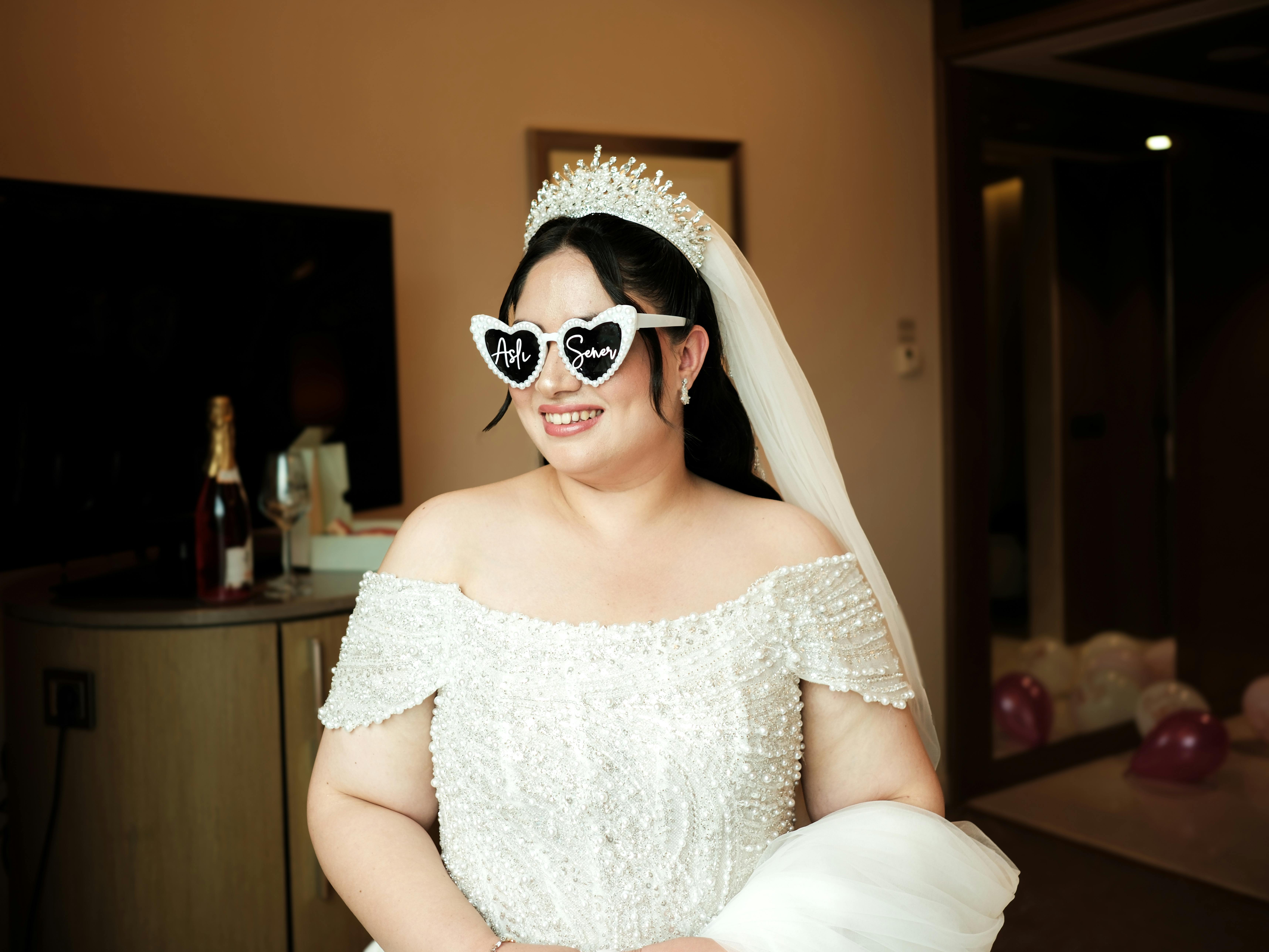 Smiling bride in elegant gown with heart sunglasses and a crown indoors.