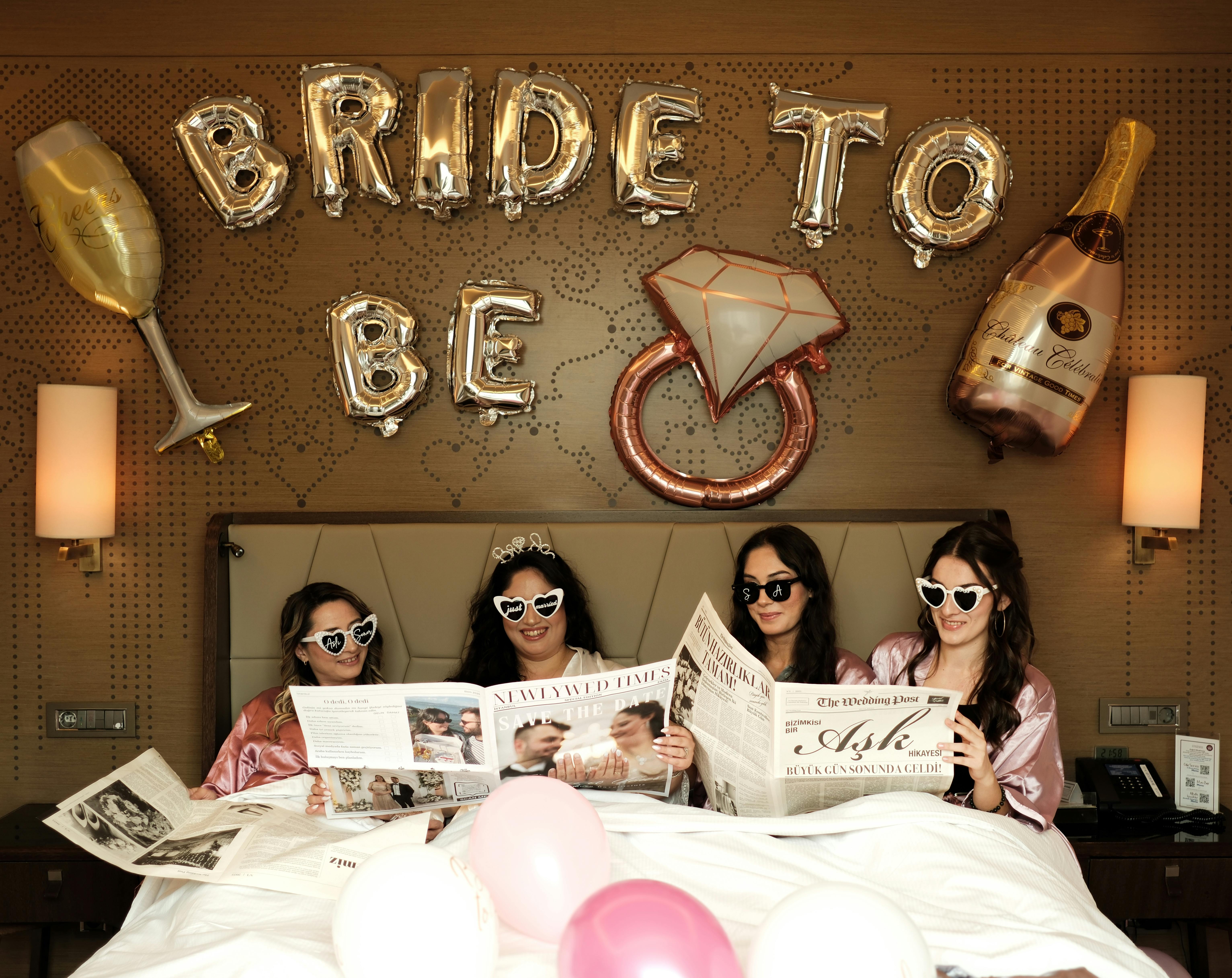 Four women celebrating a bachelorette party in a hotel room with fun props and balloons.