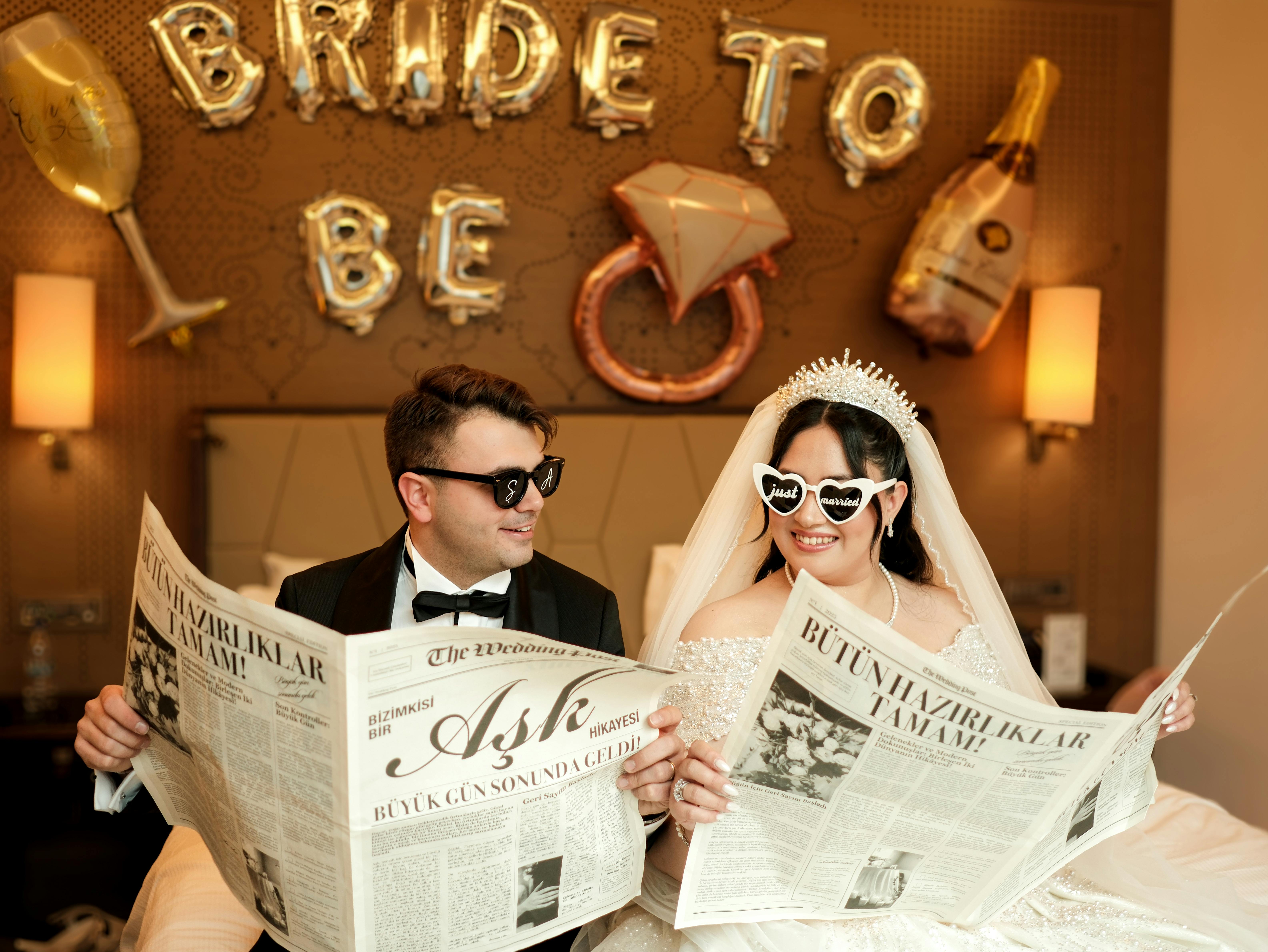 Bride and groom in sunglasses, enjoying newspapers with playful decor in hotel room.