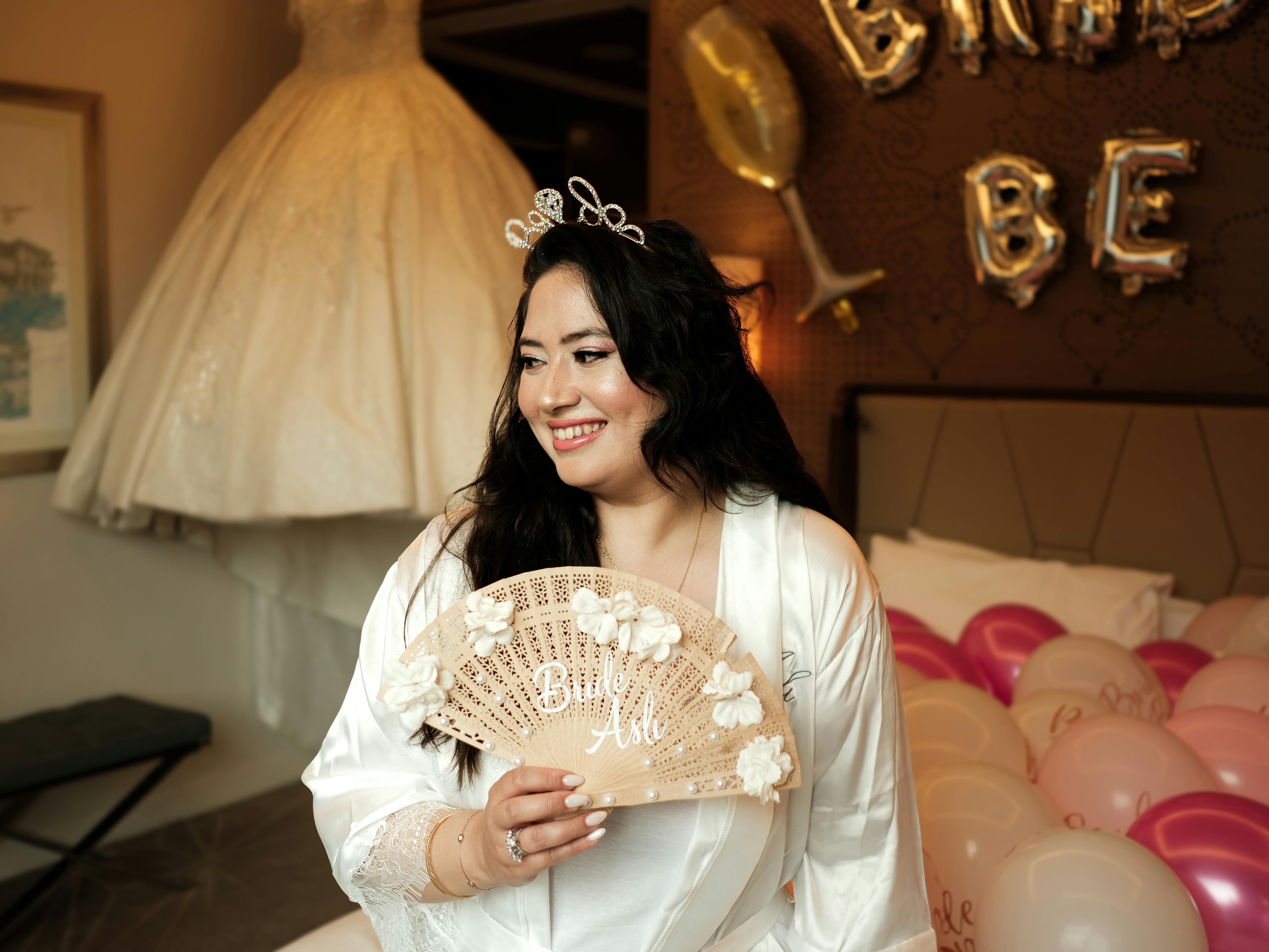 Happy bride in robe celebrating with balloons and fan in decorated room.