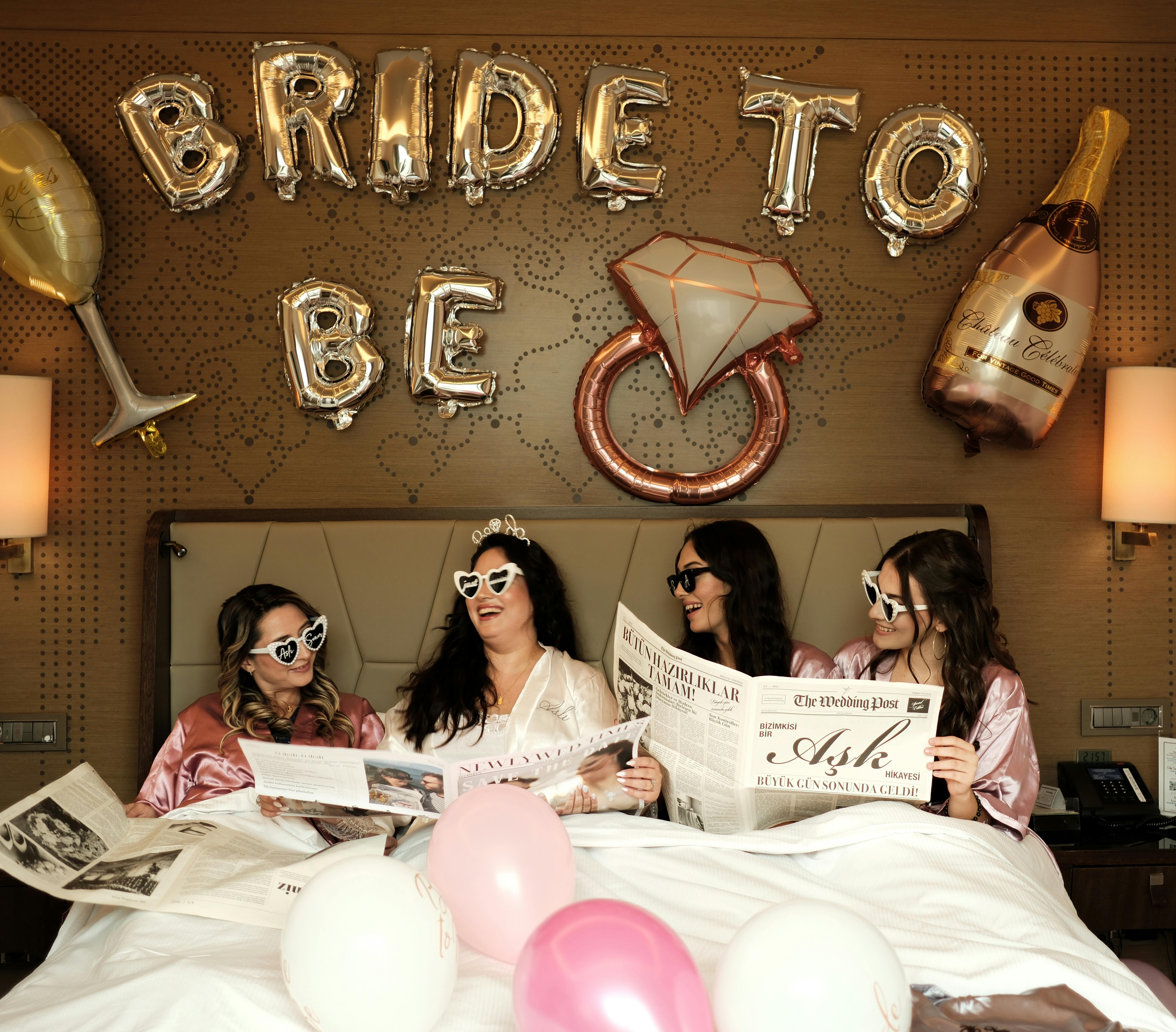 Four women celebrate a bridal shower with balloons, newspapers, and laughter.
