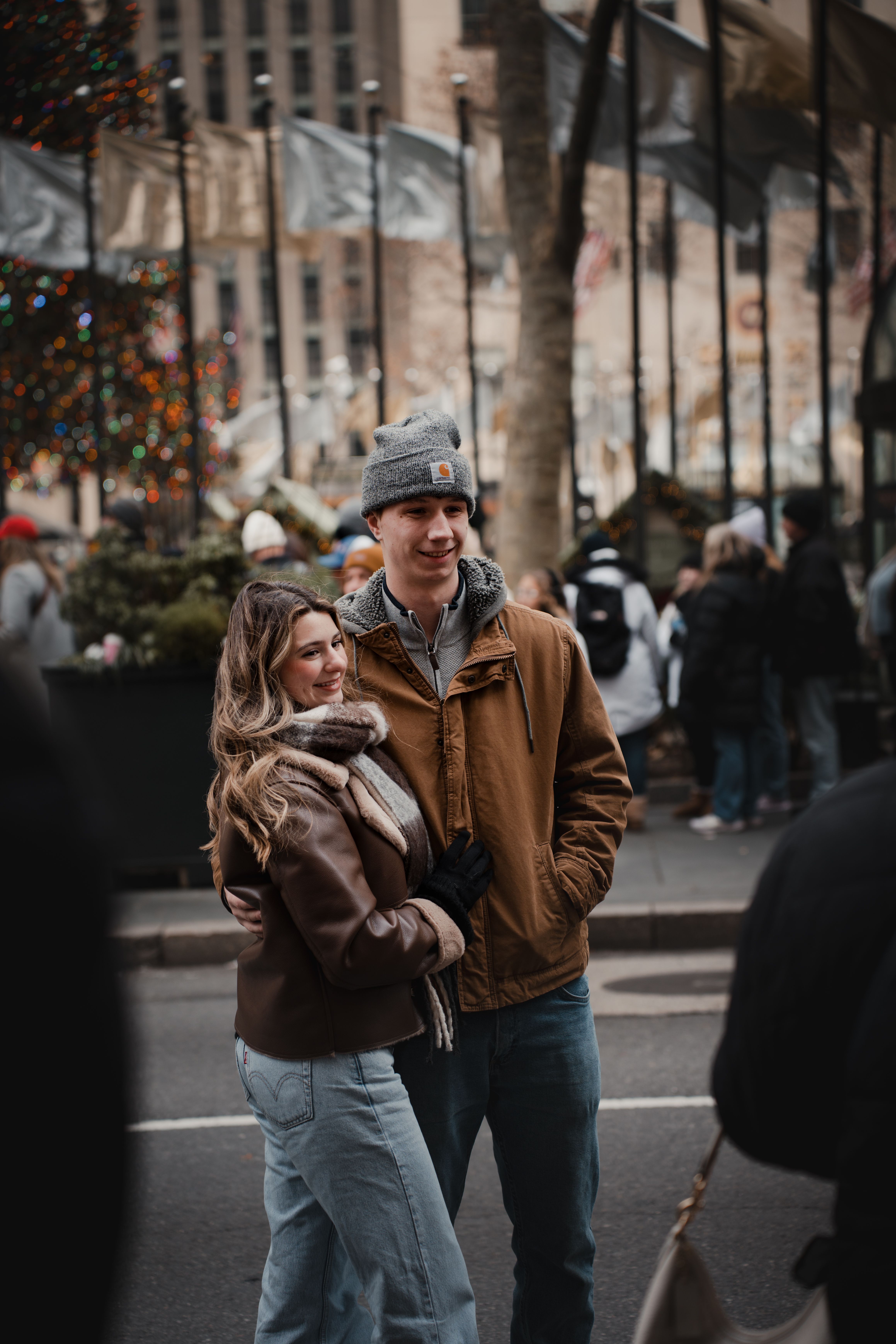 Couple embraces in a festive, urban winter setting with crowd and lights.