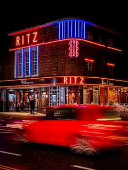Ritz at night with vibrant neon lighting, bustling street view, and red car motion blur.
