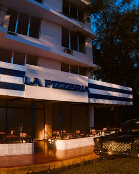 A vintage pizzeria facade illuminated by evening light with a classic car in the foreground.