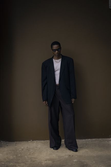 Stylish man wearing oversized suit poses in a modern, dimly-lit setting, exuding confidence.