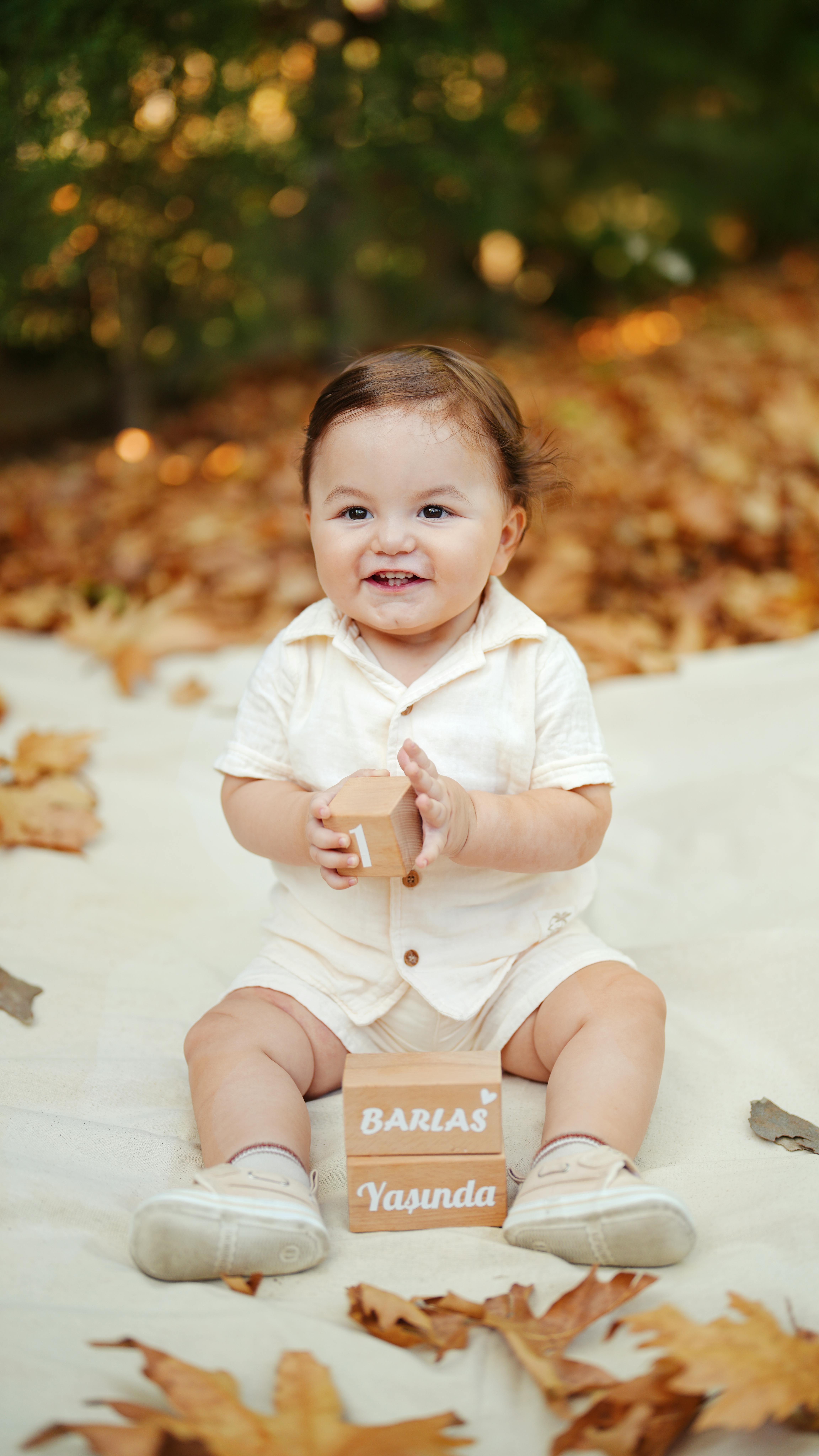 Charming baby in white outfit with a block, surrounded by autumn leaves outdoors.