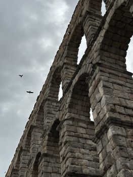 Stone arches of a Roman aqueduct reach into a cloudy sky, birds soaring.