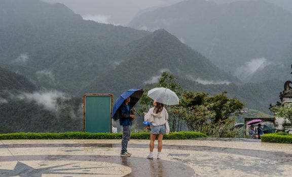 A couple stands under umbrellas on a rainy day in the mountains, surrounded by lush greenery.