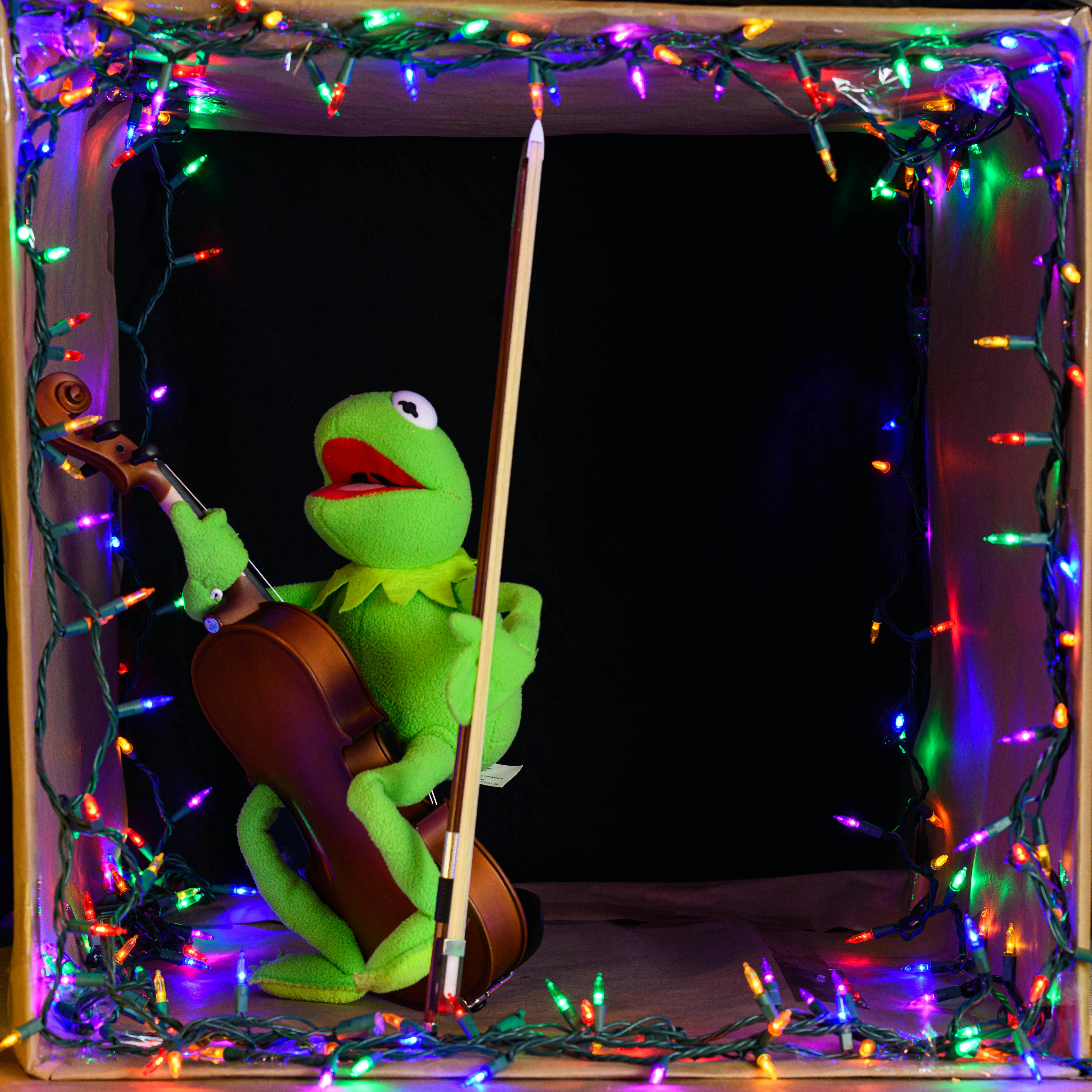 Kermit the Frog puppet plays a cello, surrounded by colorful Christmas lights, in a fun, festive setting.