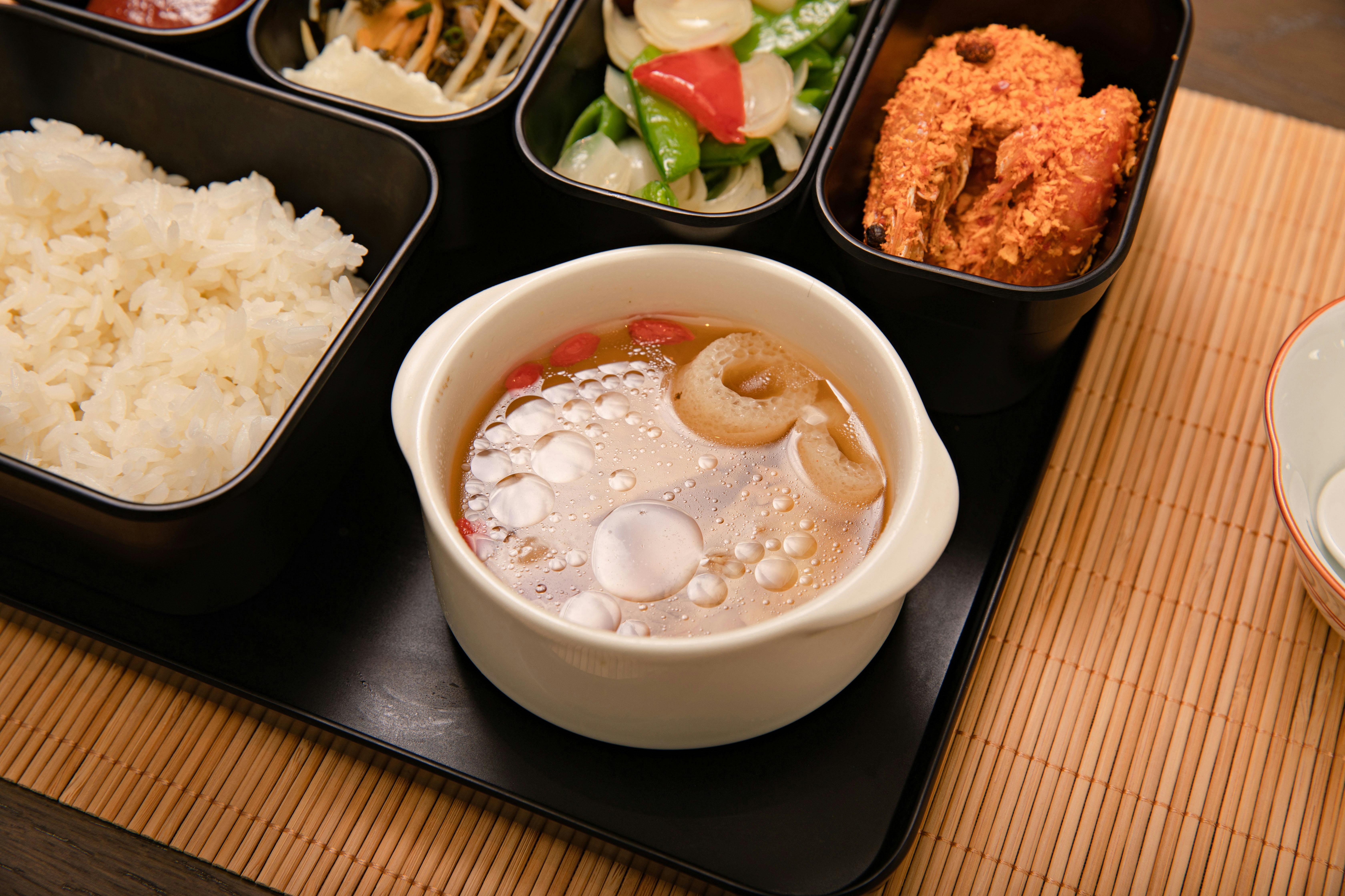 Asian bento box with rice, fried shrimp, vegetables, and flavorful soup served on a bamboo mat.