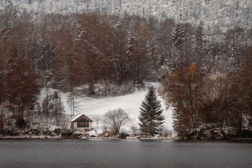 Free Serene winter scene with a cozy cabin by a snow-covered lake amidst snowy trees. Stock Photo