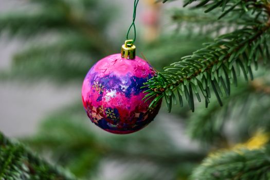 Vibrant pink and purple Christmas ornament hanging on a green evergreen branch outdoors.