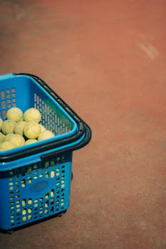 Close-up of a blue basket filled with tennis balls on a clay court.