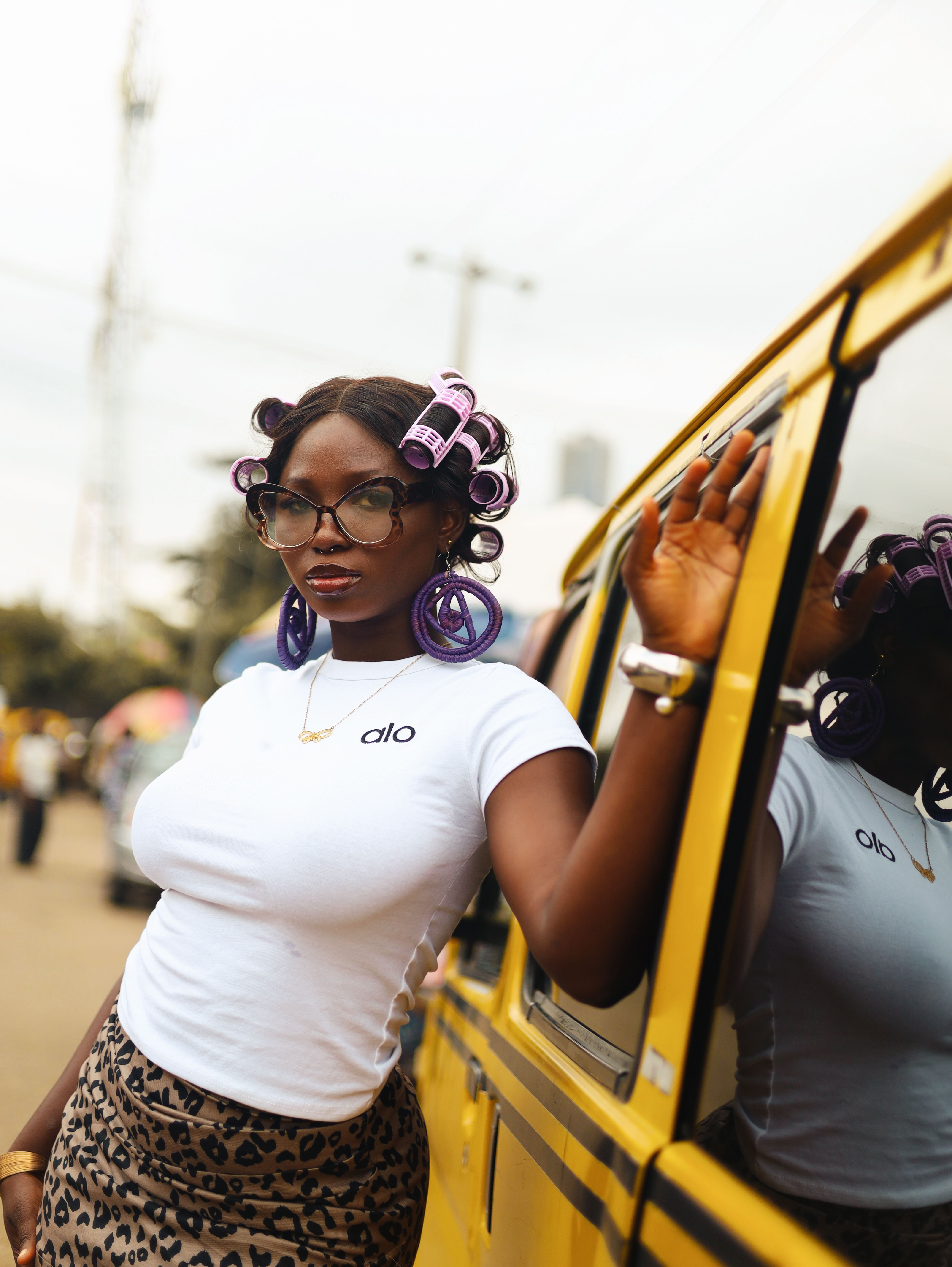 Fashionable woman with hair rollers poses by a yellow bus in a vibrant Lagos street.