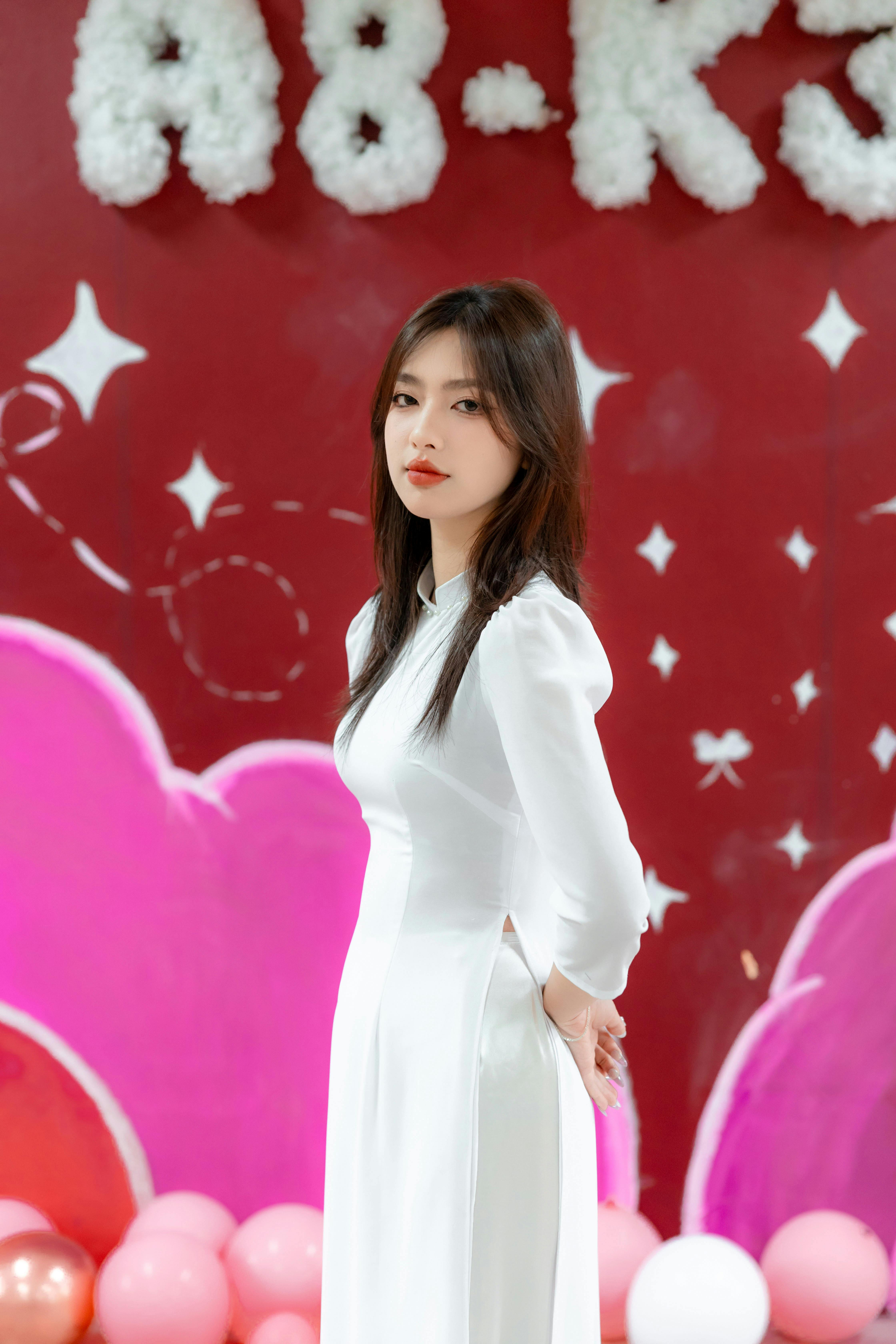 A woman in a white dress poses indoors with a colorful background and balloons.