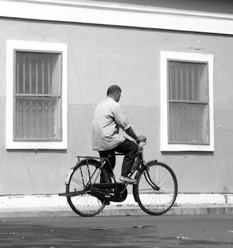 A black and white street photo featuring a man cycling past a building facade with barred windows.