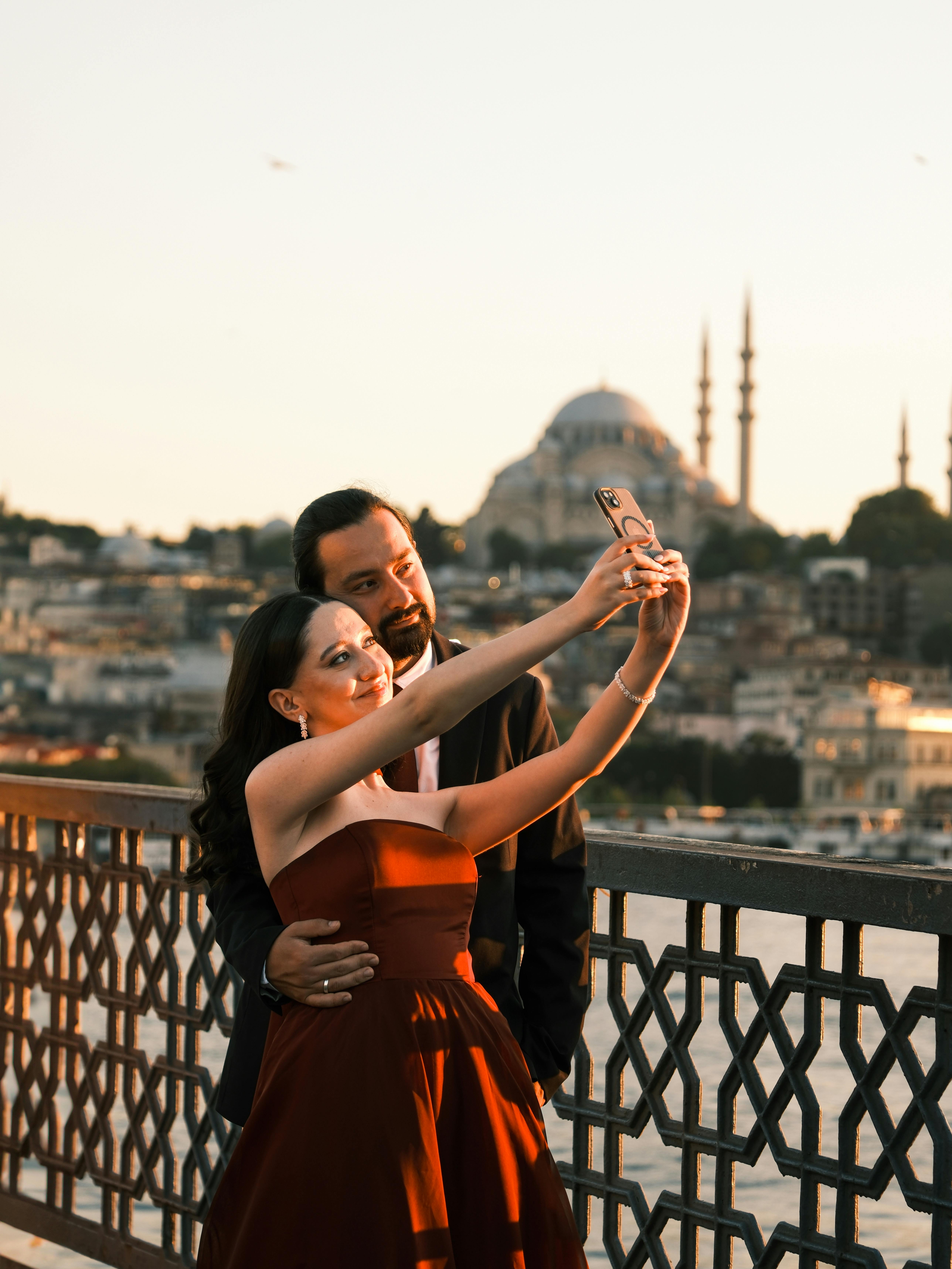 A couple takes a selfie on a bridge with the Istanbul skyline and mosque at sunset.