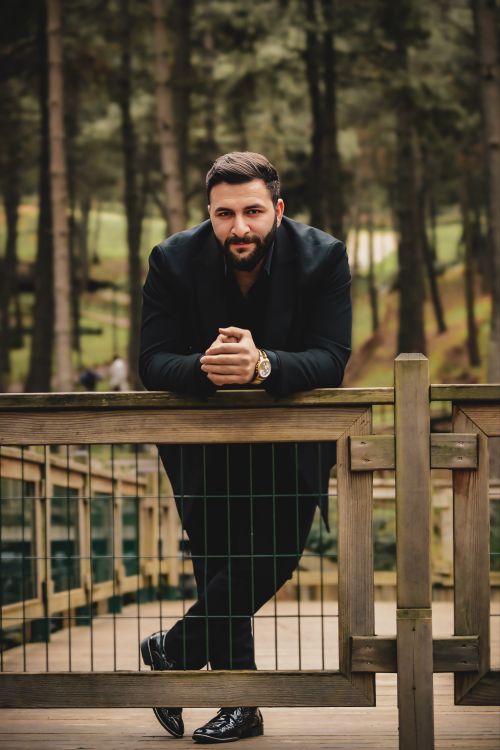 Free Stylish man in dark clothing leaning on fence in forest setting. Stock Photo