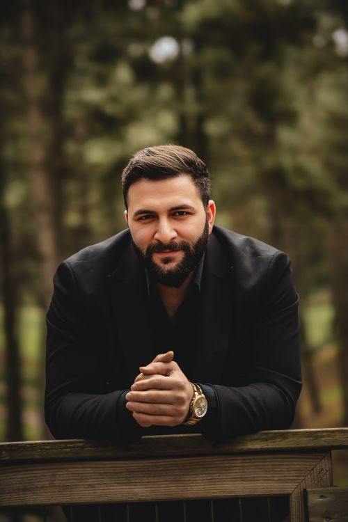 Free Charismatic man in a black suit leaning on a railing in a forest setting. Stock Photo