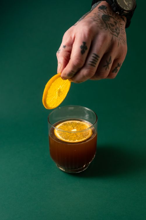 Free A tattooed hand garnishing a cocktail with an orange slice over a green background, adding flair. Stock Photo