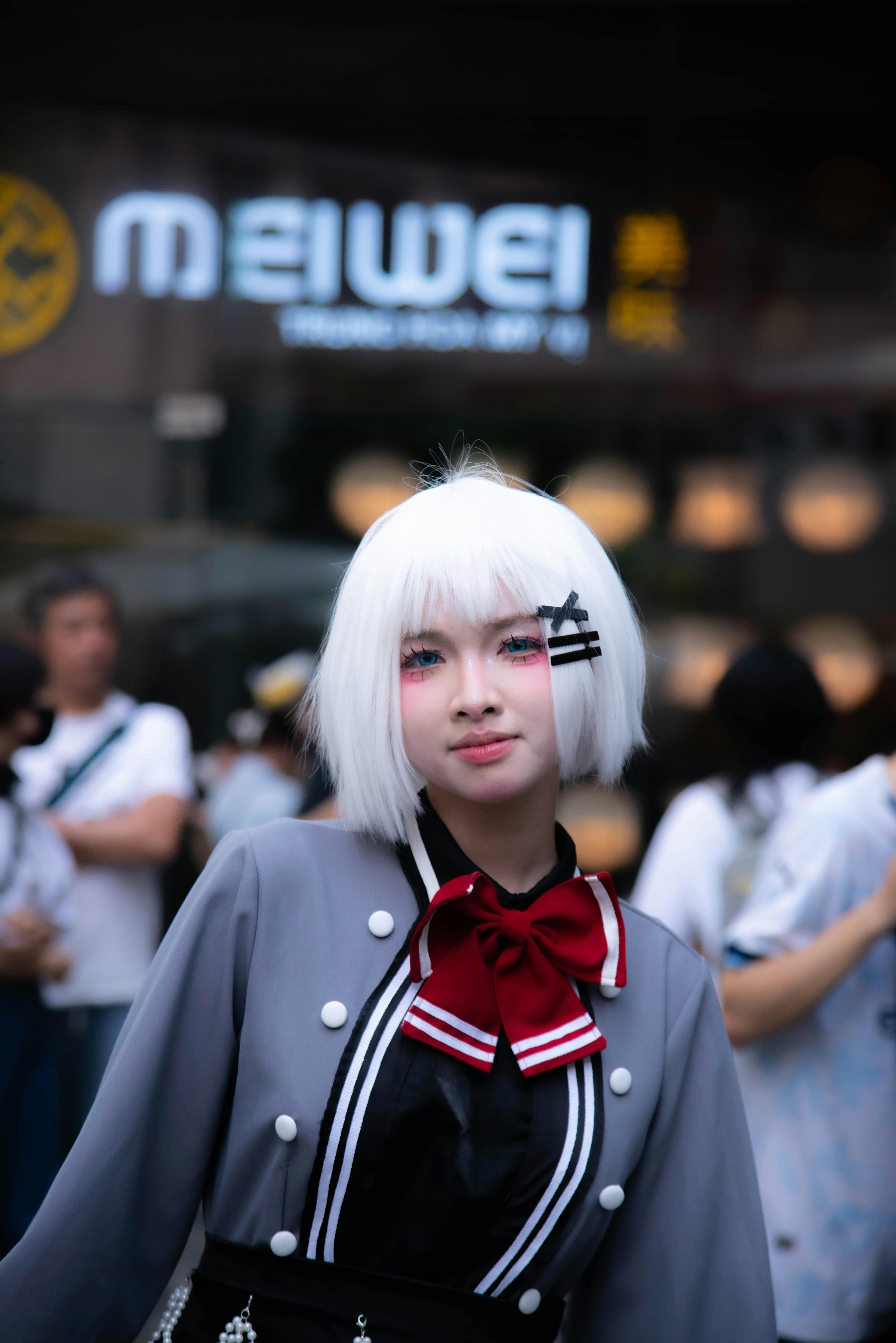 Cosplayer posing outdoors in unique costume with white hair and bow tie, urban setting in background.
