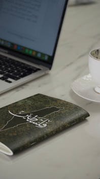 A minimalist workspace featuring a laptop, ornate notebook, and coffee cup on a marble table.