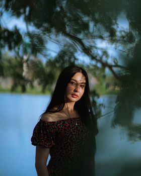A woman stands elegantly framed by leaves near a serene lake.