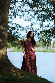 A woman in a floral dress stands by a large tree near a lake, enjoying a sunny day outdoors.