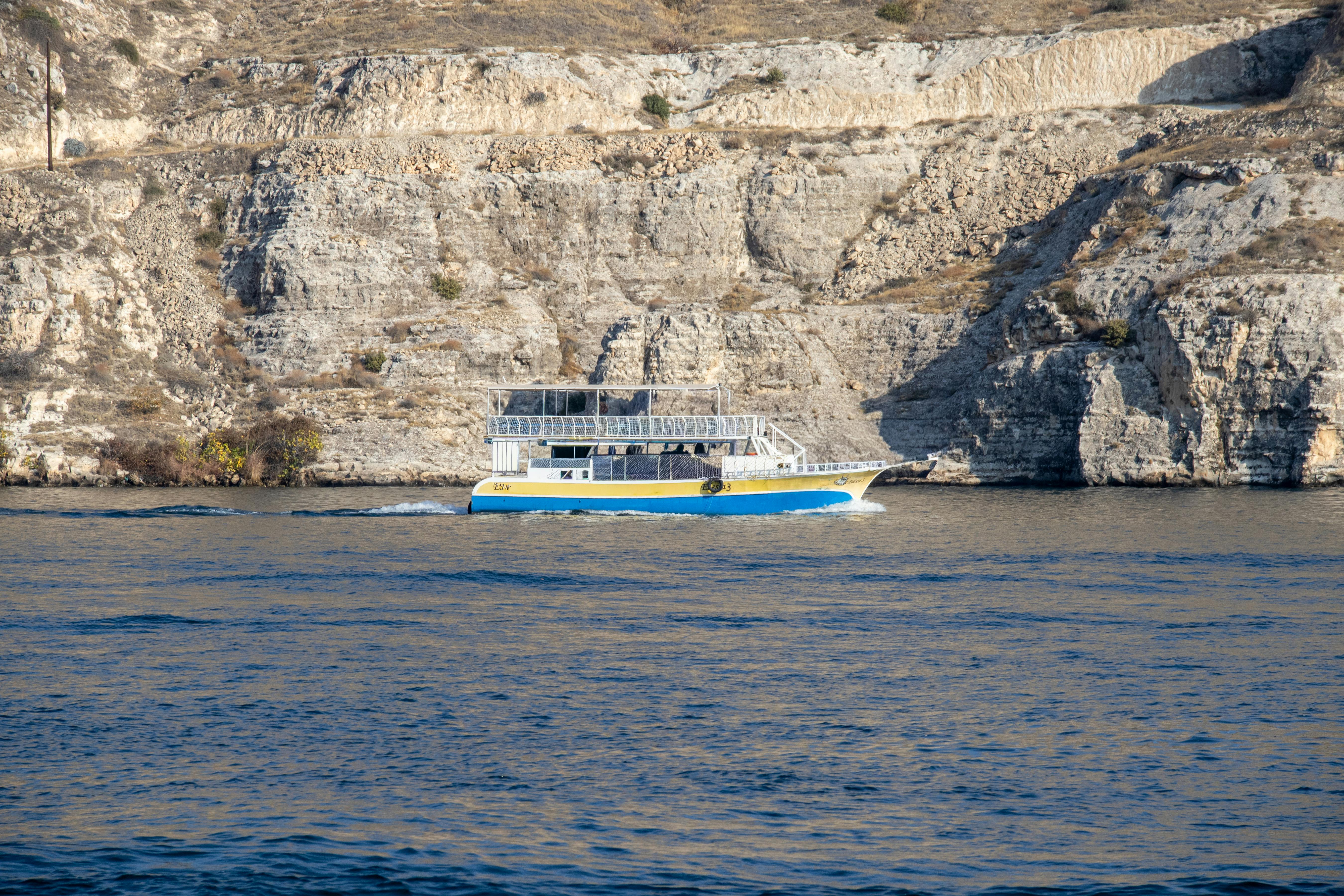 A blue and yellow boat sailing on the Euphrates River against the rocky cliffs of Halfeti, Türkiye.