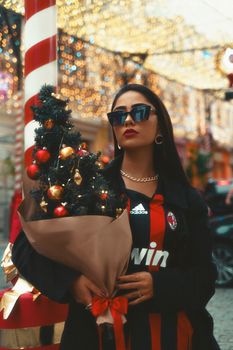 Trendy woman holding a decorated mini Christmas tree on a festive street.