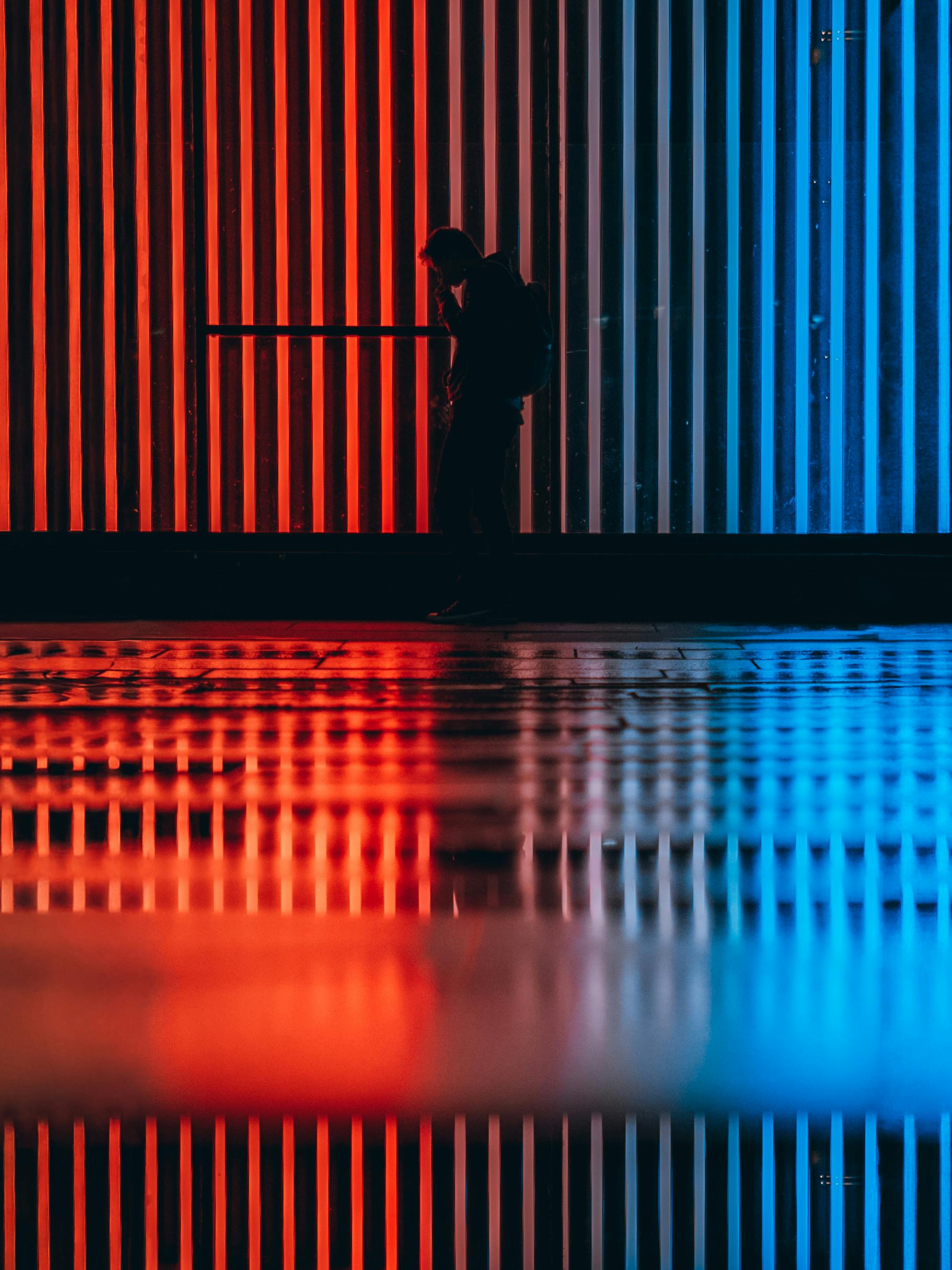 Silhouetted person stands against a striking backdrop of red and blue neon lighting with reflections.