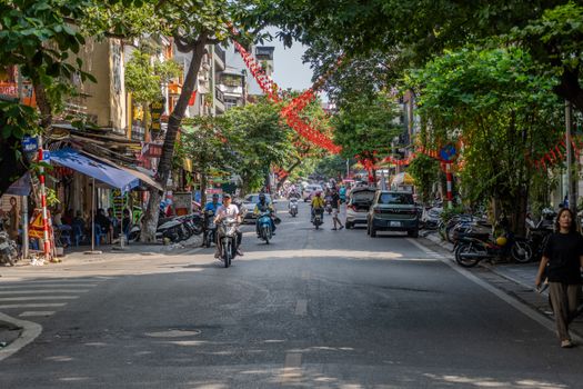 Bustling street in Hanoi with motorbikes, cars, and people under red festive decorations.